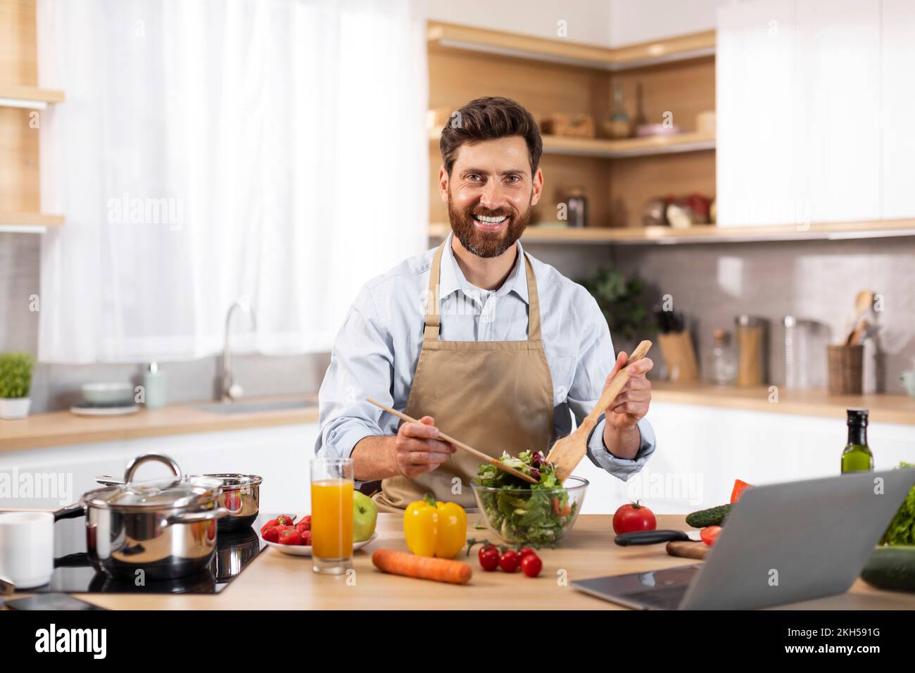 Lächelnder männlicher Koch mit europäischem Bart in der Schürze bereitet Salat in moderner Küche zu Stockfoto