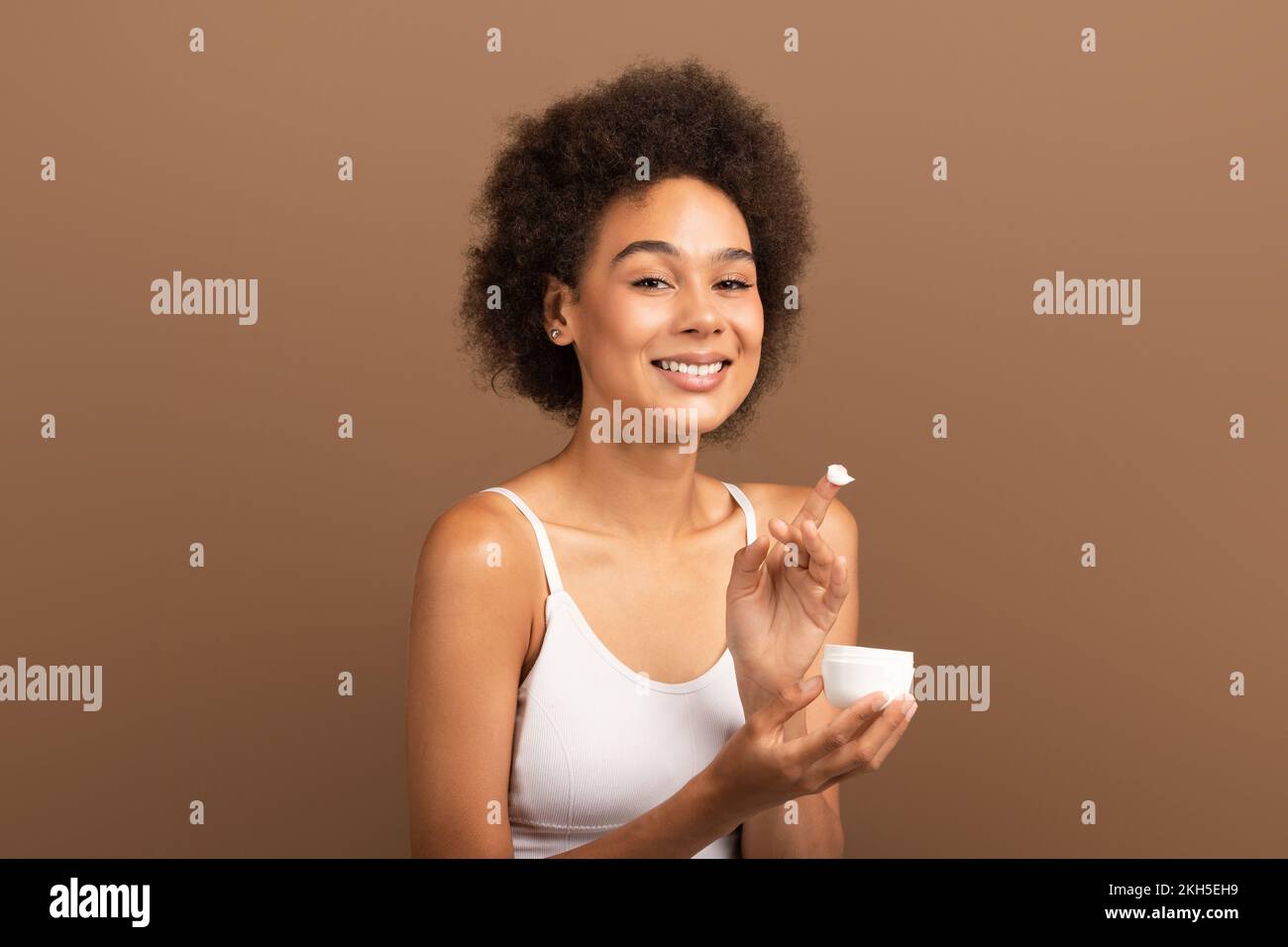 Fröhliche, afroamerikanische, lockige Millenial-Frau in weißem Glas Creme, isoliert auf braunem Hintergrund Stockfoto