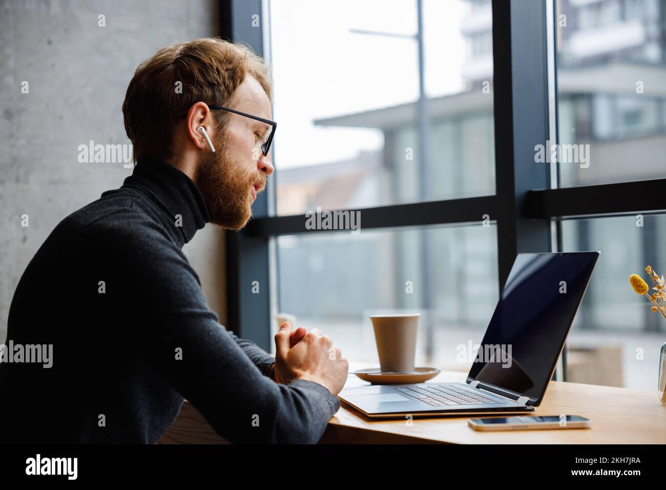 Ein junger rothaariger Typ, Programmierer oder Geschäftsmann mit Brille, sitzt in einem eleganten Café an einem Laptop und spricht über eine Webcam. Freiberufler Stockfoto