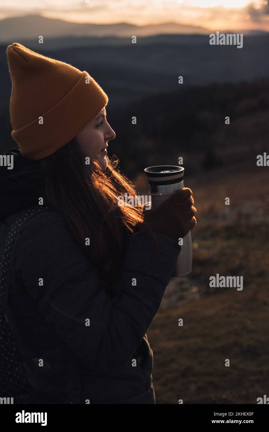 Eine junge Frau in einem orangefarbenen Beanie-Hut trinkt im Freien Tee aus einer Thermoskanne Stockfoto