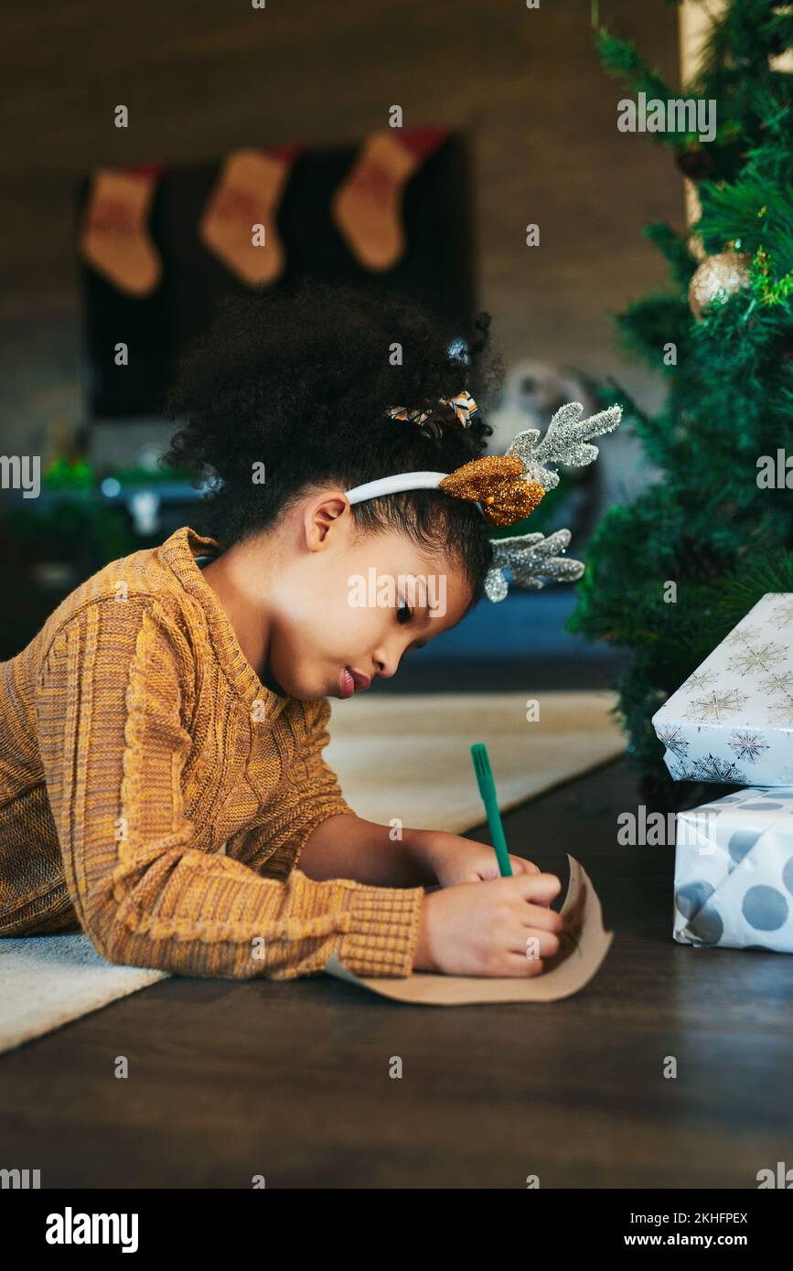 Weihnachten, Feiern und ein Mädchen, das eine Karte, Liste oder einen Urlaubsbrief auf dem Wohnzimmerboden schreibt. Idee, Wunsch und Kind mit Papier für ein Geschenk, Notiz Stockfoto