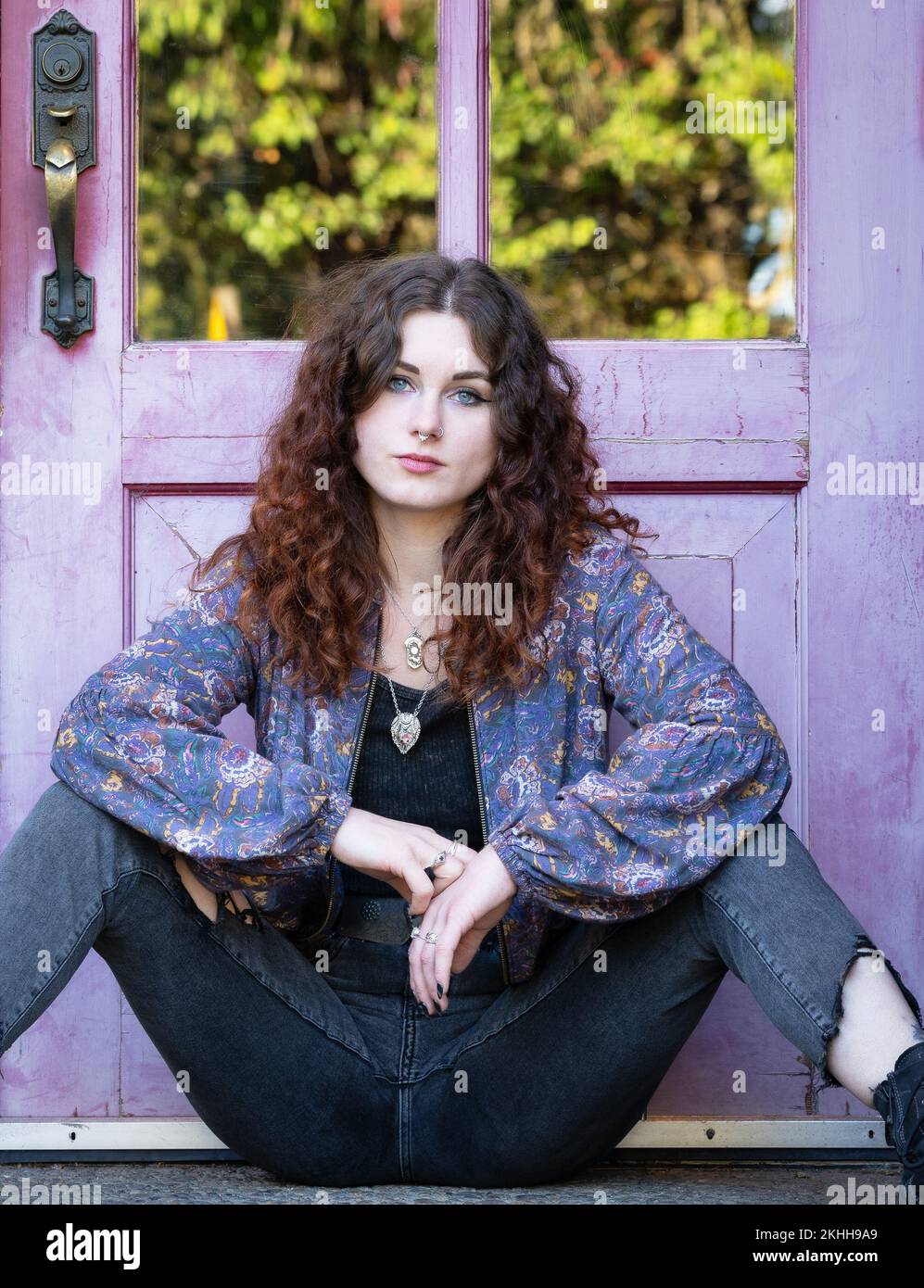 Eine Frau sitzt an einer lila Tür mit einem traurigen, nachdenklichen Gesichtsausdruck und offenen Beinen. Stockfoto