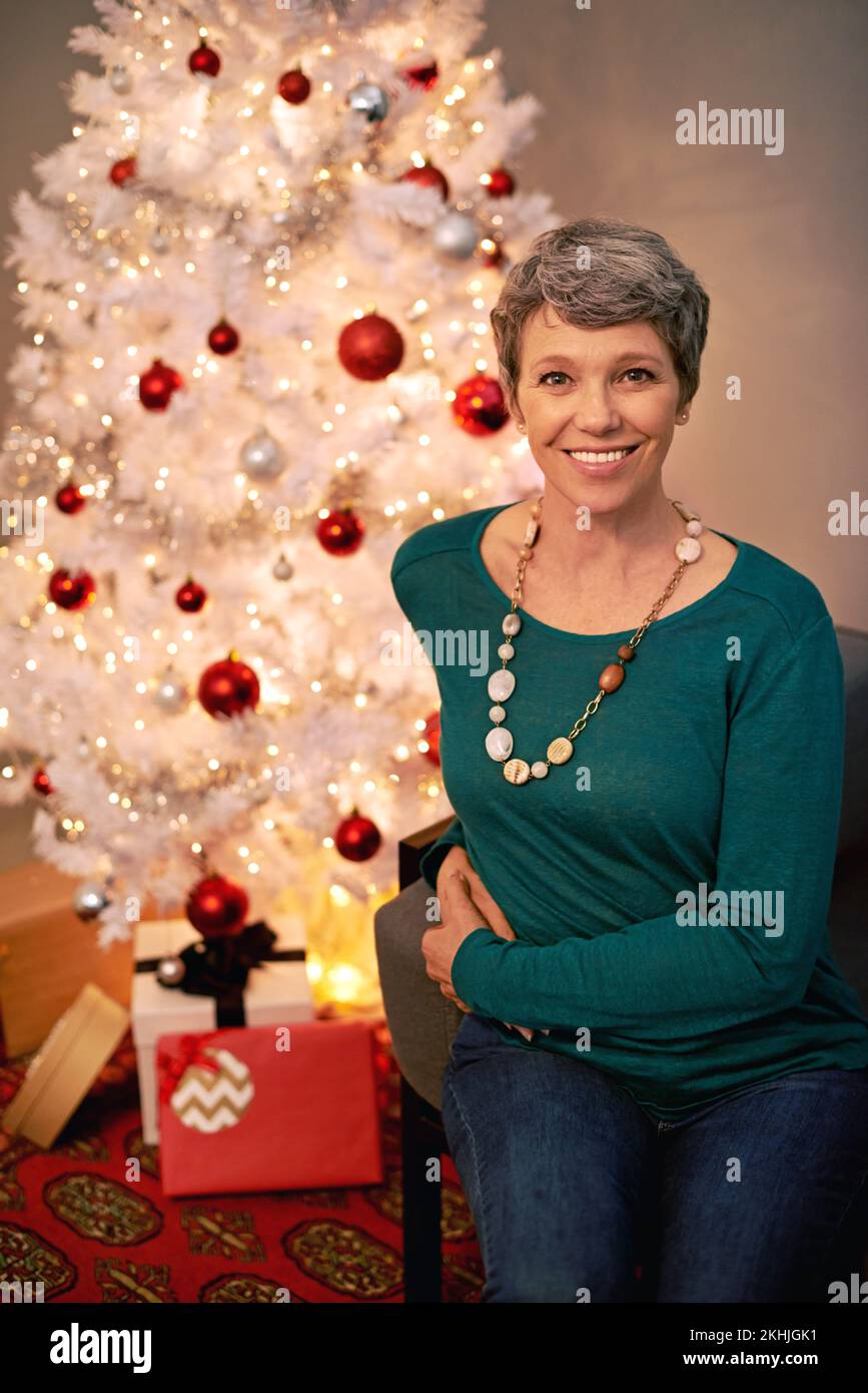 Die Jahreszeit, die alle zum Lächeln bringt. Porträt einer glücklichen, reifen Frau, die neben einem weißen Weihnachtsbaum sitzt. Stockfoto