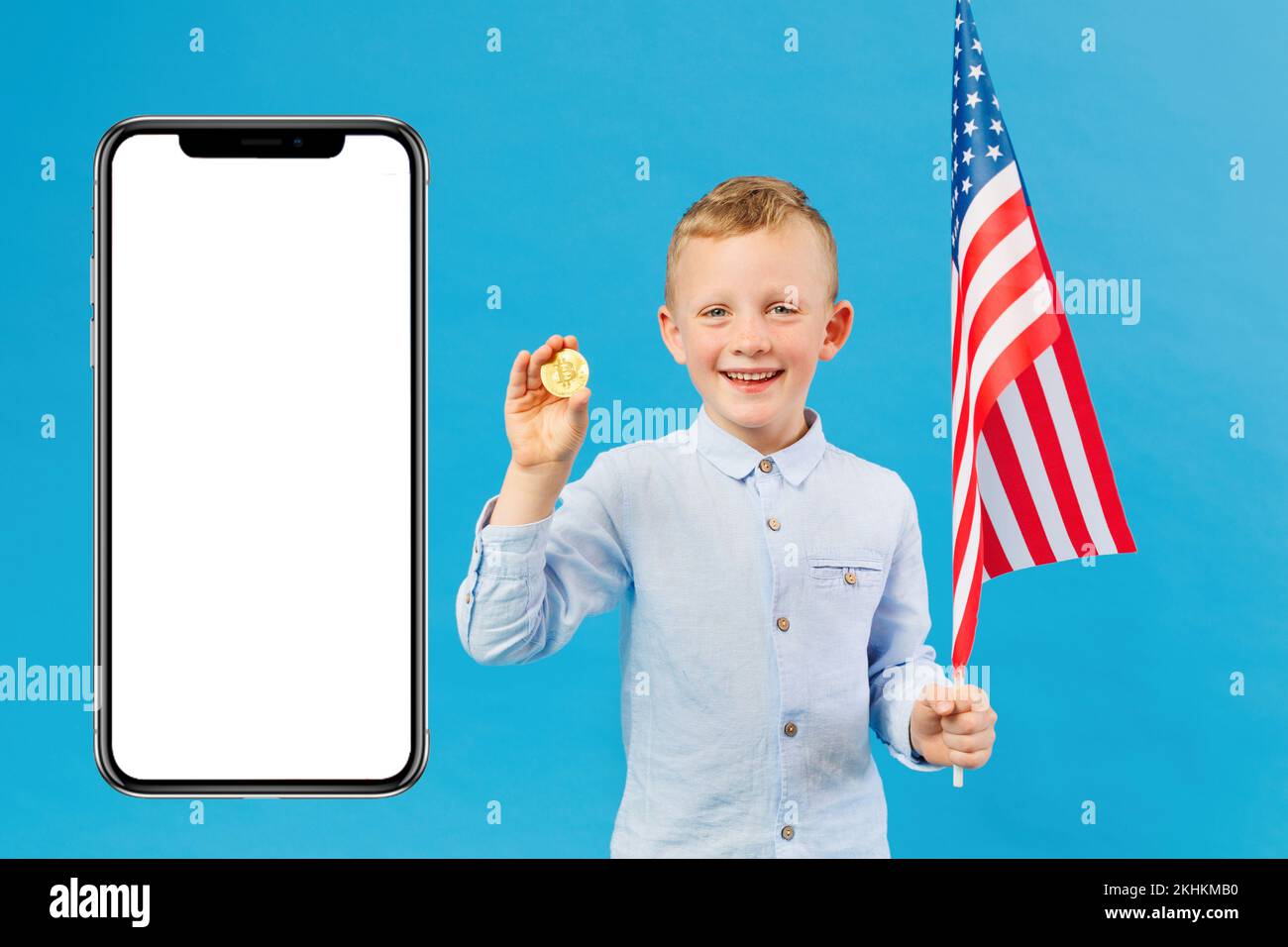 Süßer Junge mit amerikanischer Flagge und Bitcoin in einem Studio in der Halle auf blauem Hintergrund in der Nähe eines riesigen Mobiltelefons. Zukünftiges Währungskonzept Stockfoto