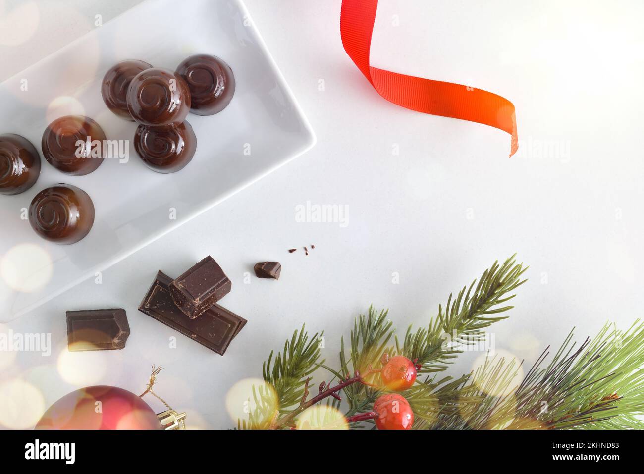 Weiße Keramikplatte gefüllt mit dunklen Schokoladenbonbons auf einem weißen Tisch mit Weihnachtsdekorationen und Portionen Schokolade als Desserts. Draufsicht. Stockfoto