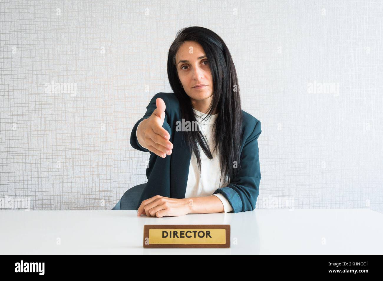 Die junge, brünette Geschäftsfrau im Büro hinter dem Schreibtisch gibt den Mitarbeitern aus der Sicht des Kunden die Hand für den Händedruck Stockfoto