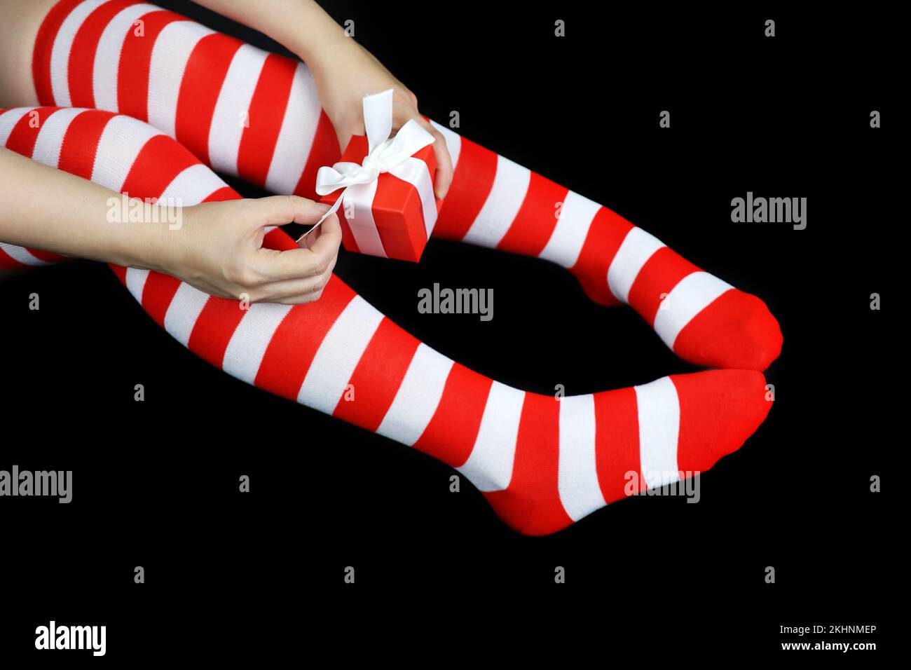 Mädchen in weihnachtlichen Kniestrümpfen mit roter Geschenkbox auf schwarzem Hintergrund. Weibliches Outfit für die Neujahrsfeier Stockfoto