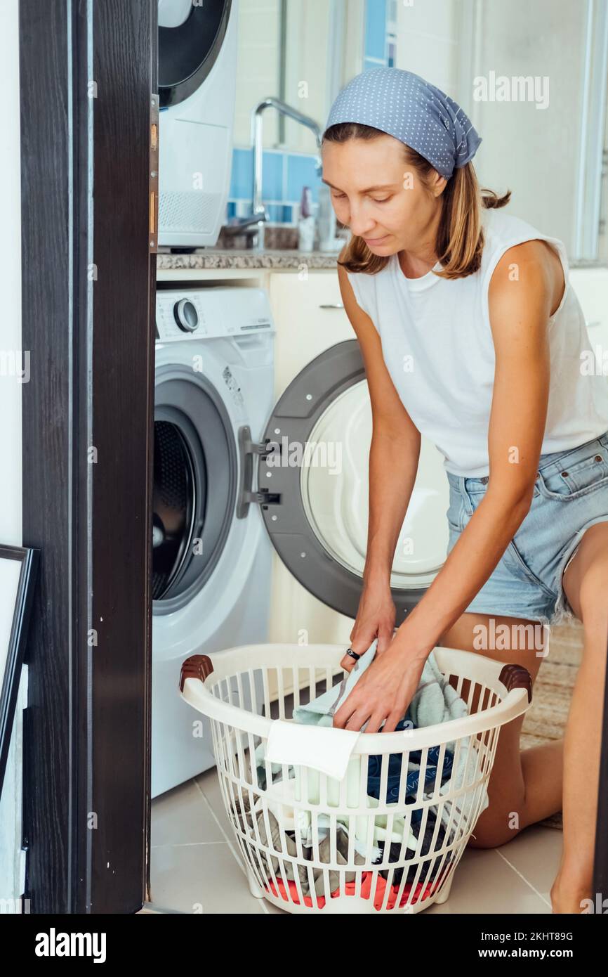 Eine junge Haushälterin sitzt vor einer Waschmaschine. Sie füllt die Waschmaschine mit dreckiger Wäsche. Stockfoto