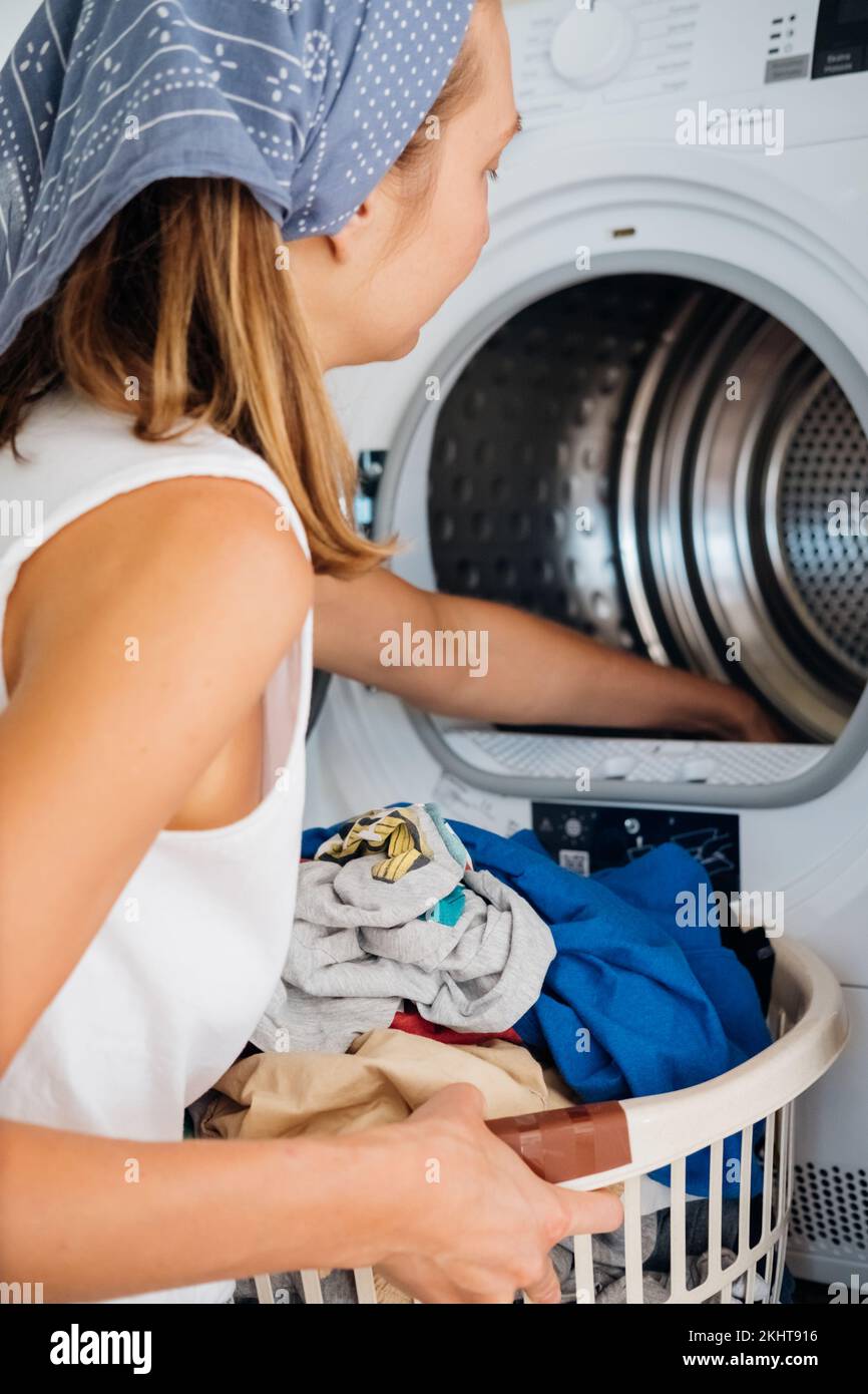Eine junge Haushälterin sitzt vor einer Waschmaschine. Sie füllt die Waschmaschine mit dreckiger Wäsche. Stockfoto