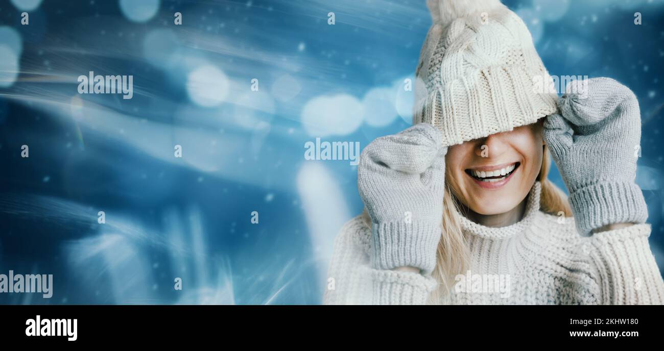 Lächelnde Frau in warmen Kleidern auf blauem Winterhintergrund. Versteck das Gesicht unter dem Strickmütze aus Wolle. Banner mit Kopierbereich Stockfoto