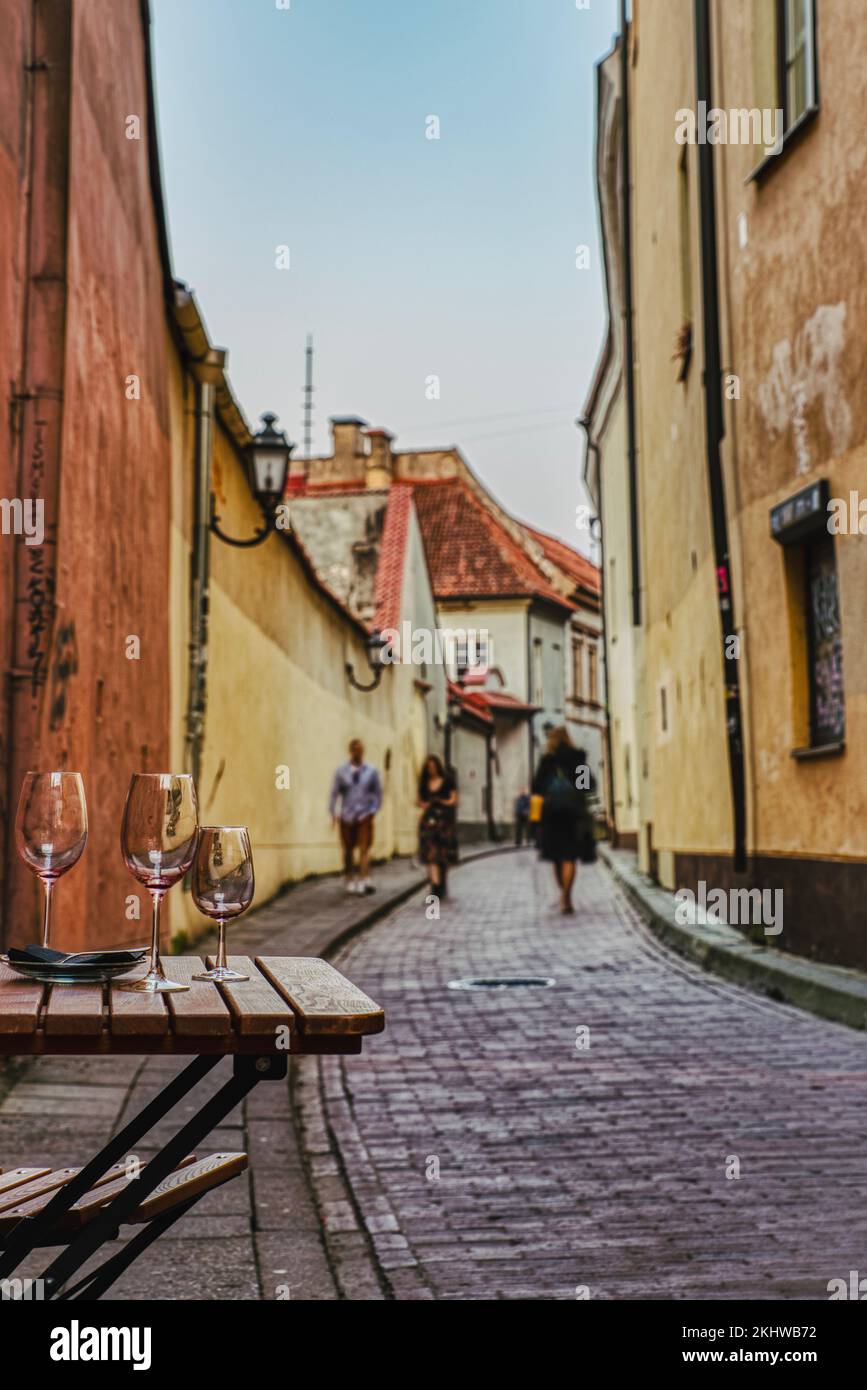 Gepflasterte gemütliche Gasse in der historischen Altstadt von Vilnius mit Schwerpunkt auf einem rustikalen Tisch an der Seite mit einem leeren Glas Wein, während verschwommene Menschen gehen. Litauen Stockfoto