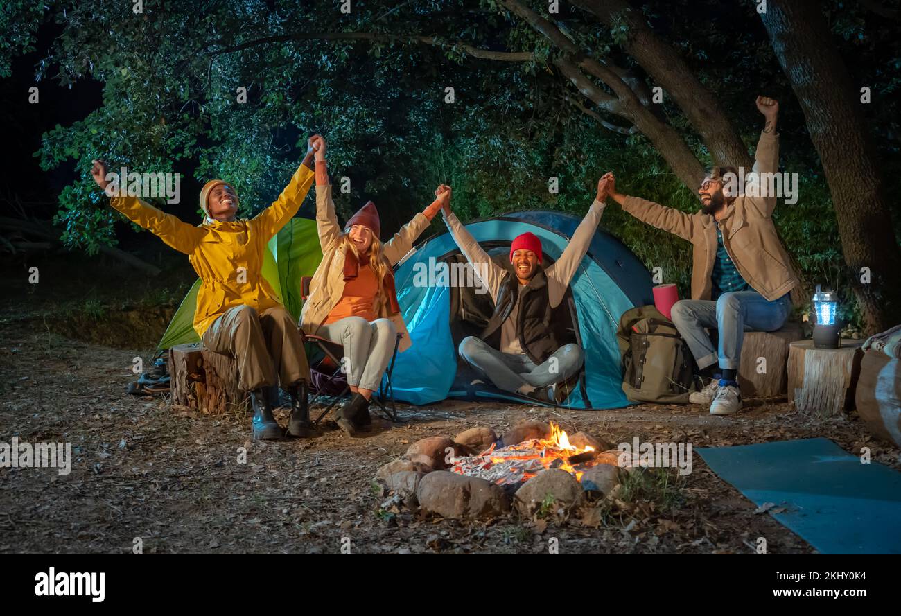 Fröhliche Freunde feiern und gemeinsam mit den Armen das Lagerfeuer auf dem Camping erobern Stockfoto
