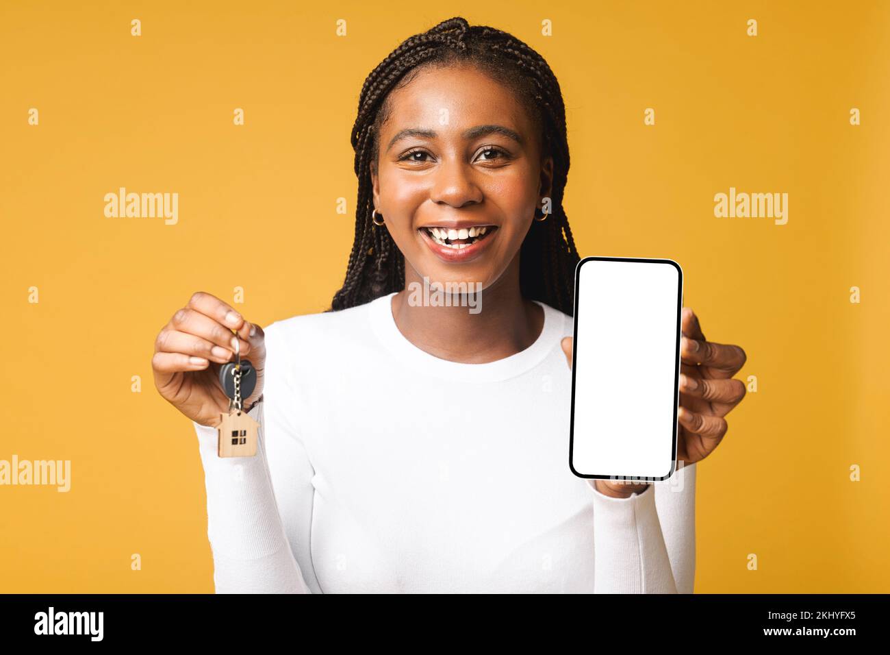 Porträt einer bezaubernden, multiethnischen Frau, die den Hausschlüssel hält und ein Smartphone auf gelbem Hintergrund im Studio mit Kopierbereich präsentiert Stockfoto