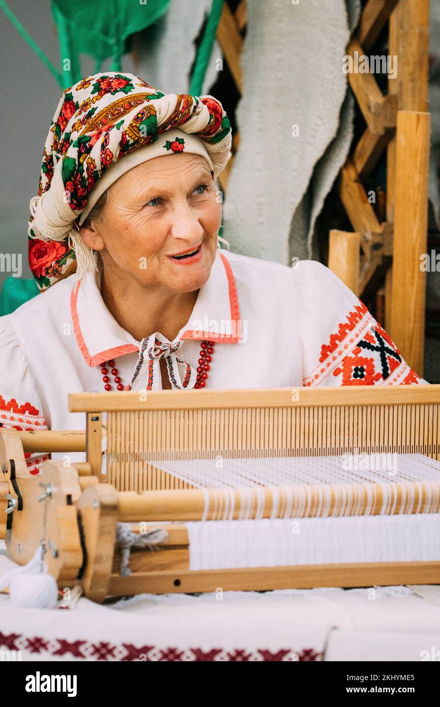 Ältere weibliche Weberin in traditionell bestickter belarussischer Kleidung hinter Einer Webmaschine. Volksfest In Minsk, Belarus. Stockfoto
