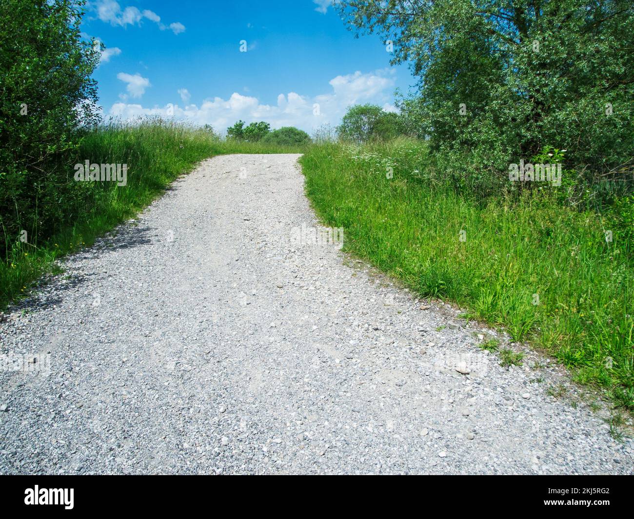 Sehen Sie eine bergauf kieselige Straße in einer hügeligen Landschaft, umgeben von Gras und kleinen Bäumen vor einem blauen Sommerhimmel mit kleinen weißen Wolken. Stockfoto