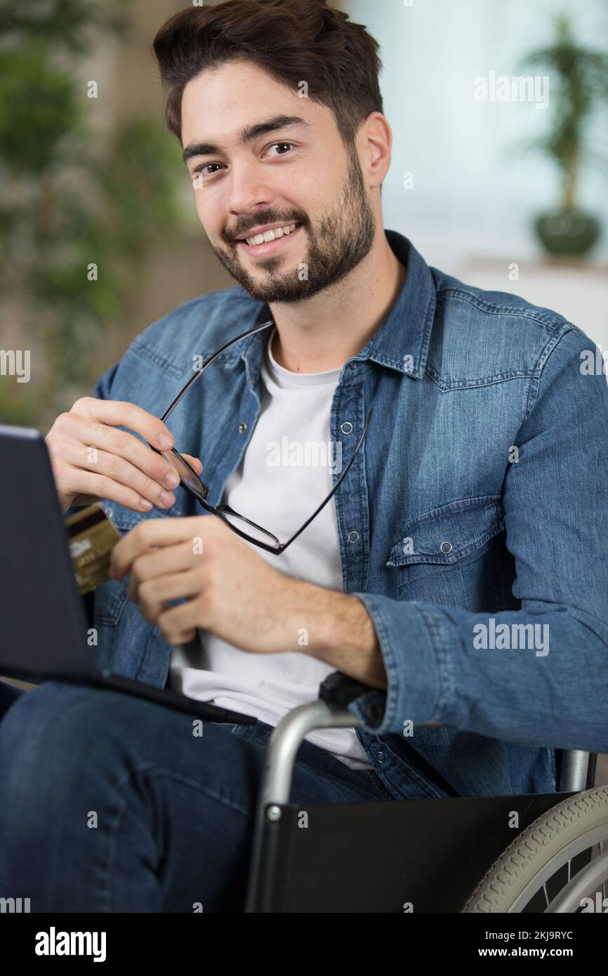 Ein Mann im Rollstuhl, der einen Laptop benutzt und bankcard in der Hand hat Stockfoto