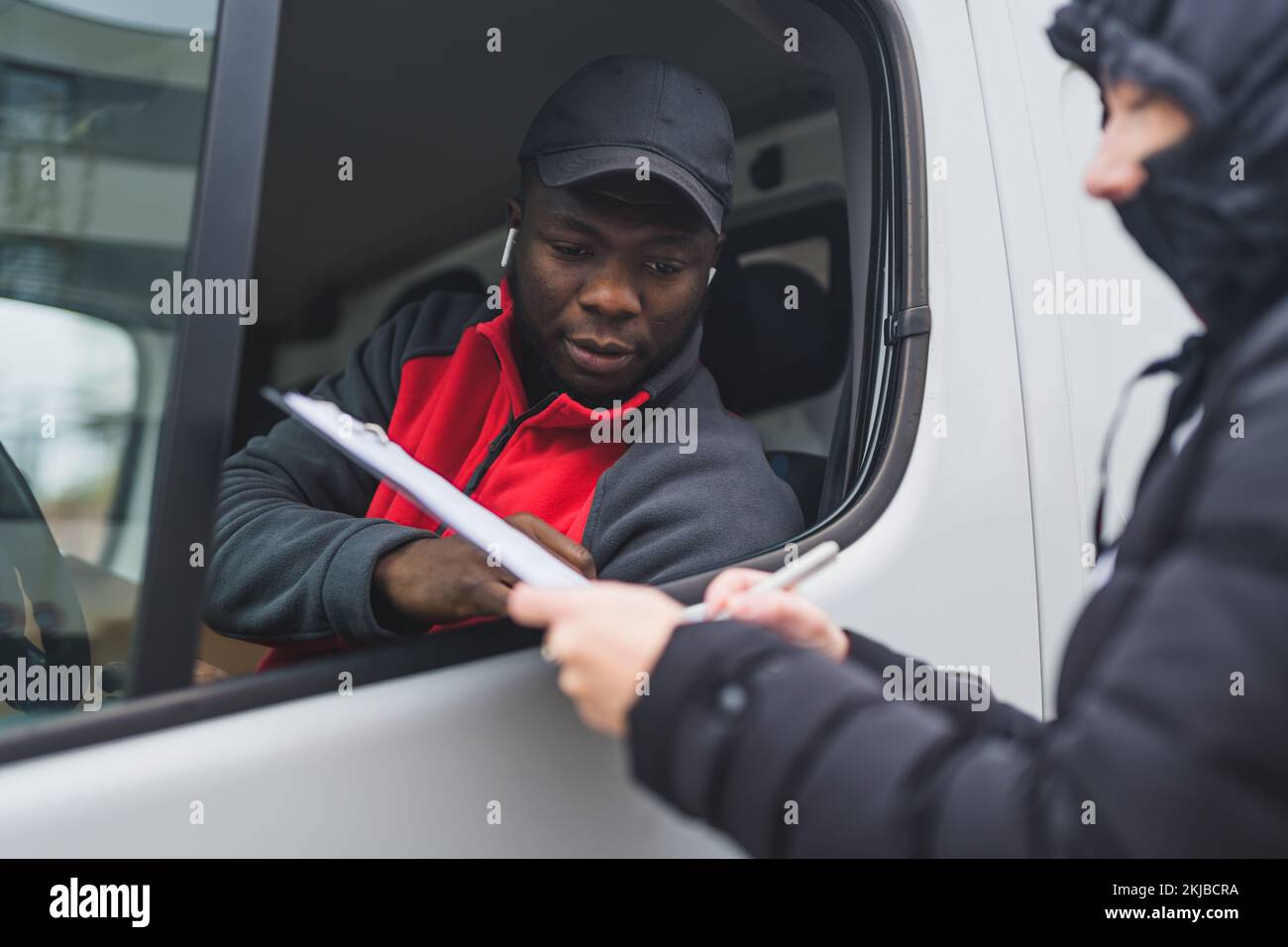 Eine Person in einer Puffy Jacke, die Papiere unterschreibt, hielt den schwarzen jungen erwachsenen Liefermann mit rotem Pullover aus dem Fenster des weißen Lieferwagens. Horizontalschuss. Hochwertiges Foto Stockfoto