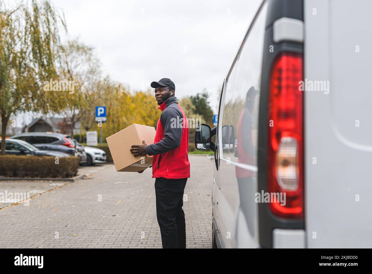 Schwarzer junger Erwachsenenlieferant in rotem Pullover, der beim weißen Lieferwagen steht und nach einem Haus sucht, an das das Paket geliefert werden soll. Horizontalschuss im Freien. Hochwertiges Foto Stockfoto