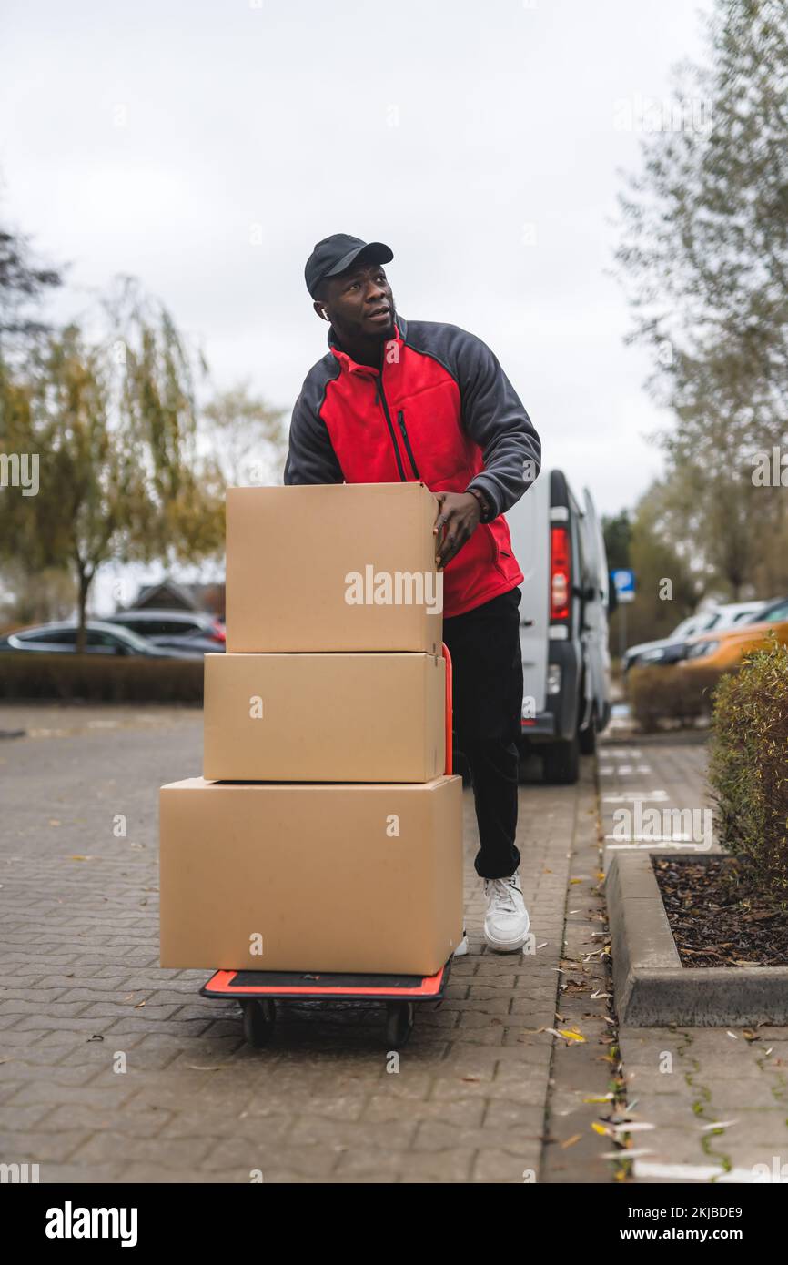 Junger erwachsener schwarzer Lieferjunge, der einen roten Uniformpullover trägt, der Pakete auf einem Trolley stapelt. Vertikales Außenfoto. Hochwertiges Foto Stockfoto