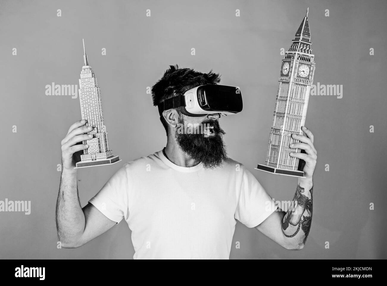 Mann mit Bart in VR-Brille, grüner Hintergrund. Kerl in VR-Brille hält Big Ben und Empire State Building. 3D-Designkonzept. Mann auf glückliches Gesicht Studie Stockfoto