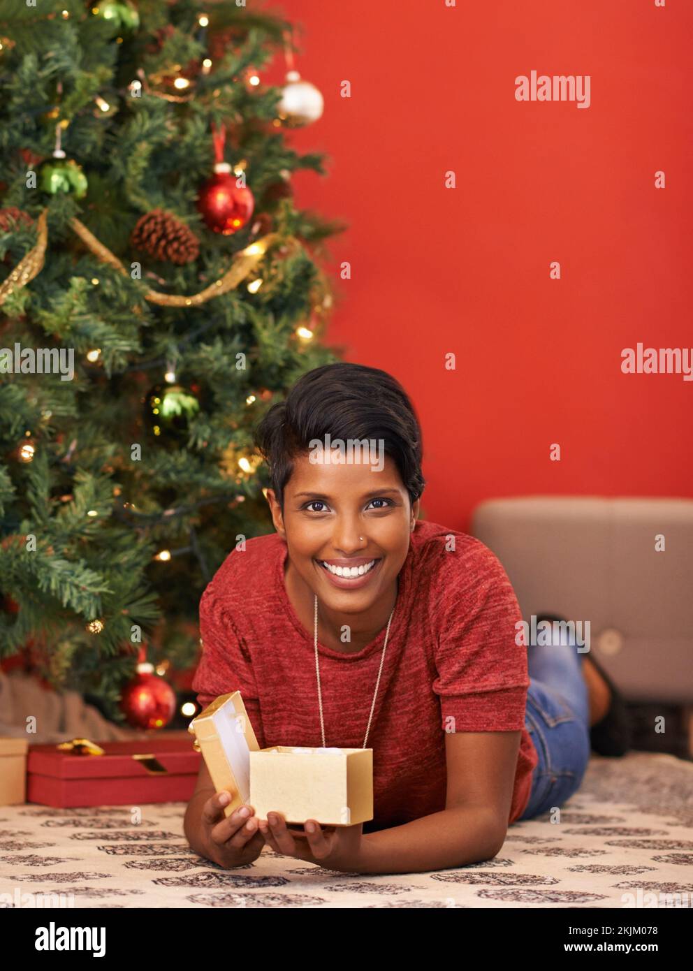 Ich öffne immer ein Geschenk am Heiligabend. Eine attraktive junge Frau, die ihr Geschenk am Weihnachtsbaum öffnet. Stockfoto