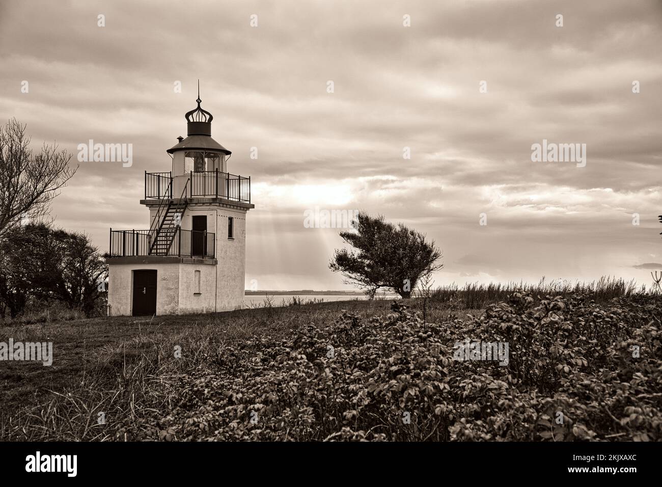 Leuchtturm in Sepia, Spodsbjerg Fyr in Huntsted an der Küste Dänemarks. Die Sonne scheint durch die Wolken. Wiese mit Bäumen. PH-Wert Querformat Stockfoto