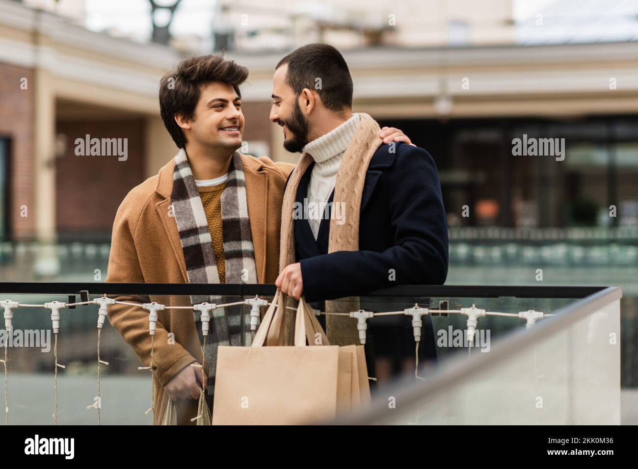 Fröhliche Schwule in trendigen Mänteln und Schals, die Einkaufstüten in der Hand halten und sich auf der Straße ansehen, Stockbild Stockfoto