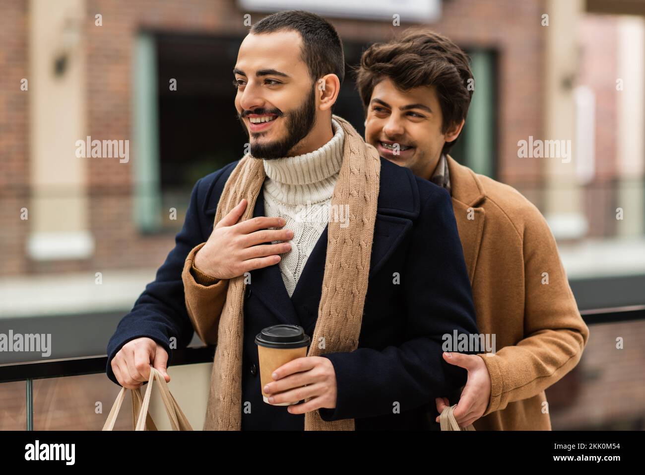 Bärtige Schwule in Mantel und Schal stehen mit einem Getränk zum Mitnehmen neben ihrem Freund, umarmen ihn draußen, Börsenbild Stockfoto
