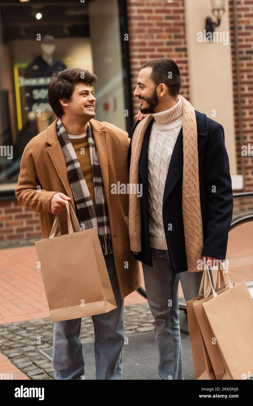 ein schwules Paar in stilvollem Outfit, das Einkaufstaschen in der Hand hält und sich auf der Straße anlächelt, Stockbild Stockfoto