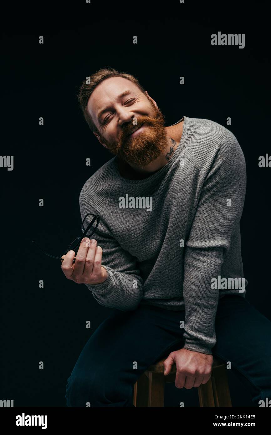 Lächelnder bärtiger Mann im Jumper, der eine Brille hält, während er auf dem Stuhl sitzt, isoliert auf schwarzem Stockbild Stockfoto