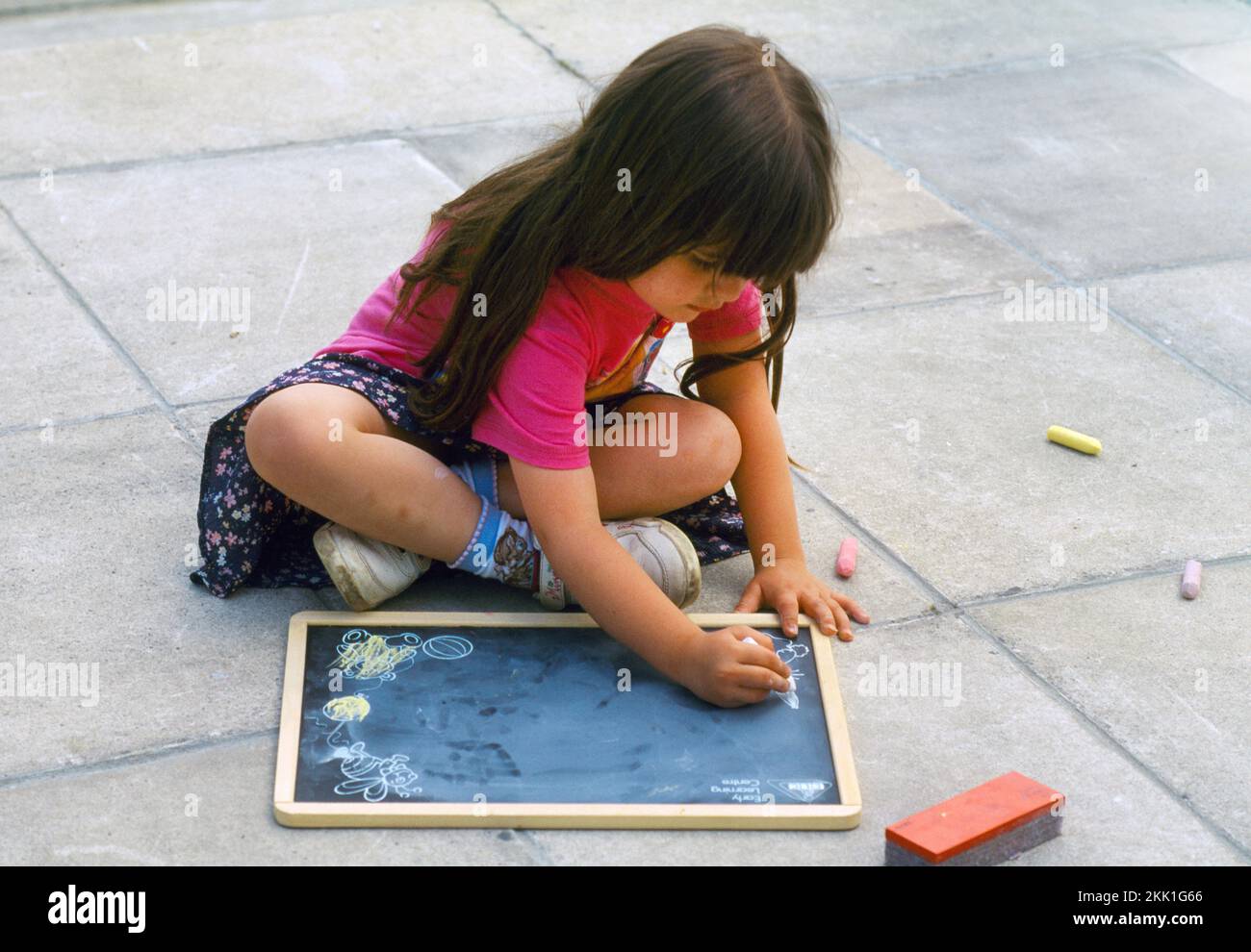 3-jähriges Mädchen, das im Garden Surrey England mit Kreide auf einer Tafel sitzt Stockfoto