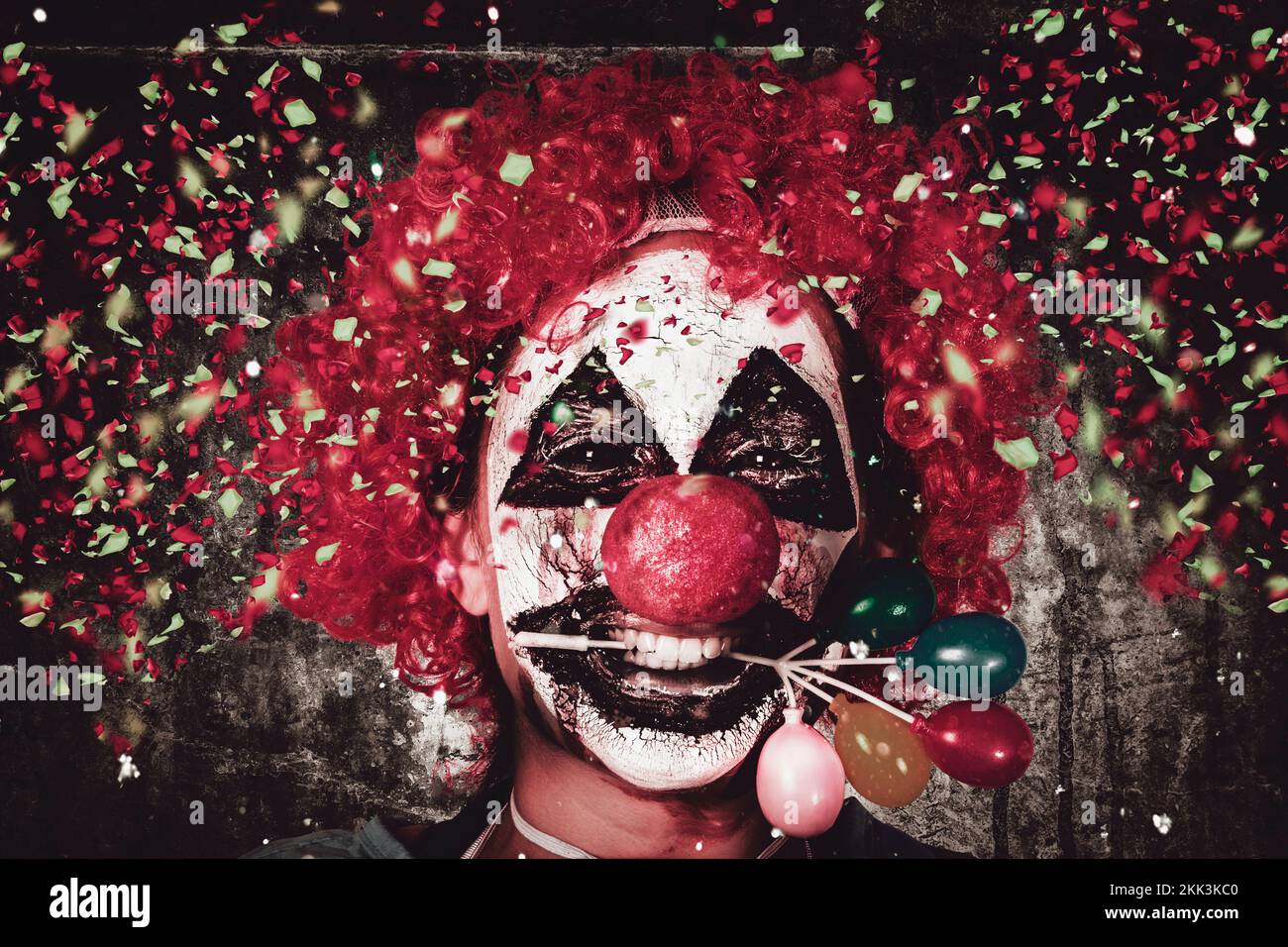 Horizontales Nahaufnahme-Porträt auf dem Gesicht eines verrückten Faschings-Clowns, der unter einem Konfetti-Sturz eine Ballonkuchendekoration im Mund hält. Wir Feiern Den Hallow Stockfoto