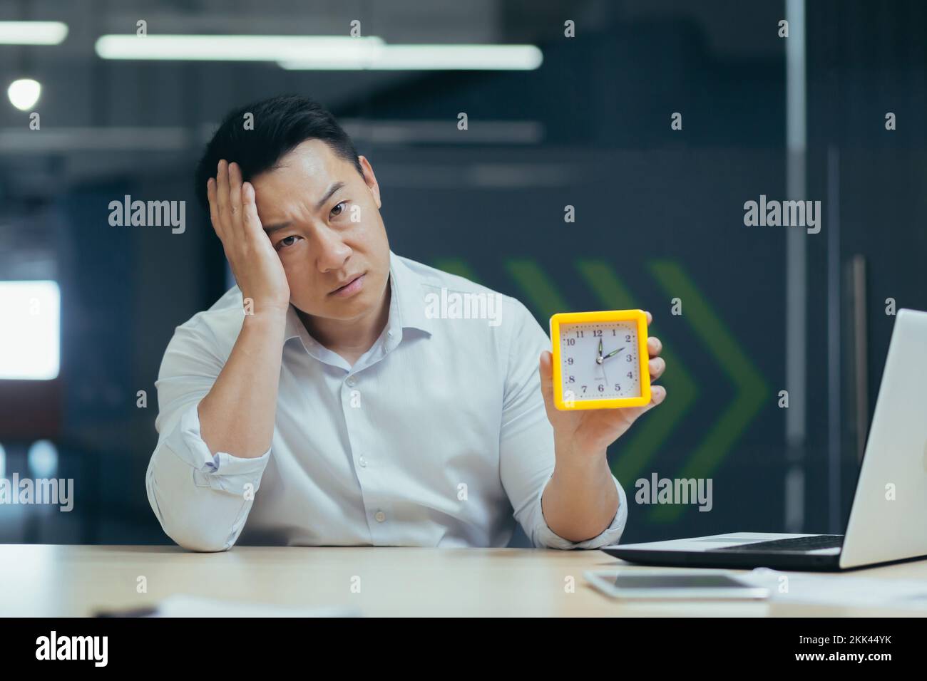 Erschöpfung bei der Arbeit, Burnout. Ein müder junger asiatischer Geschäftsmann, der im Büro sitzt und eine Uhr in der Hand hält. Schläft ein, legt seinen Kopf auf seine Hand. Warten auf das Ende des Arbeitstages. Stockfoto