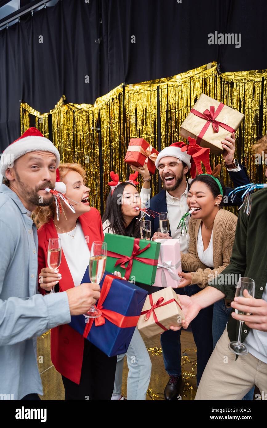 Fröhliche, gemischtrassige Geschäftsleute mit Partyhörnern, die im Büro Geschenke und Champagner halten Stockfoto