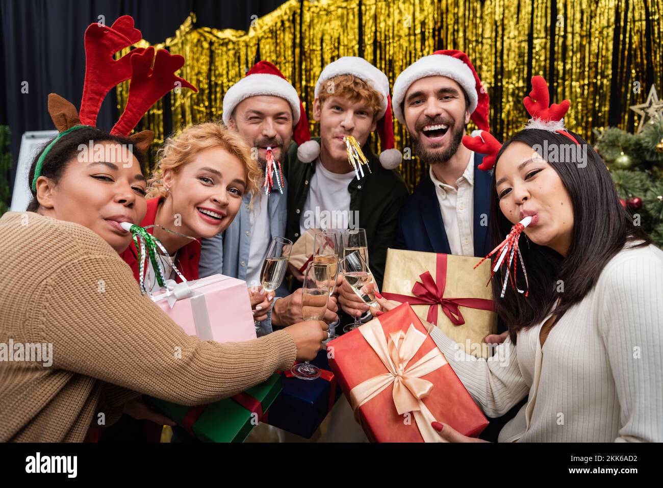 Multikulturelle Geschäftsleute mit Partyhörnern, die Geschenke halten und während der Silvesterparty Champagner klingeln Stockfoto