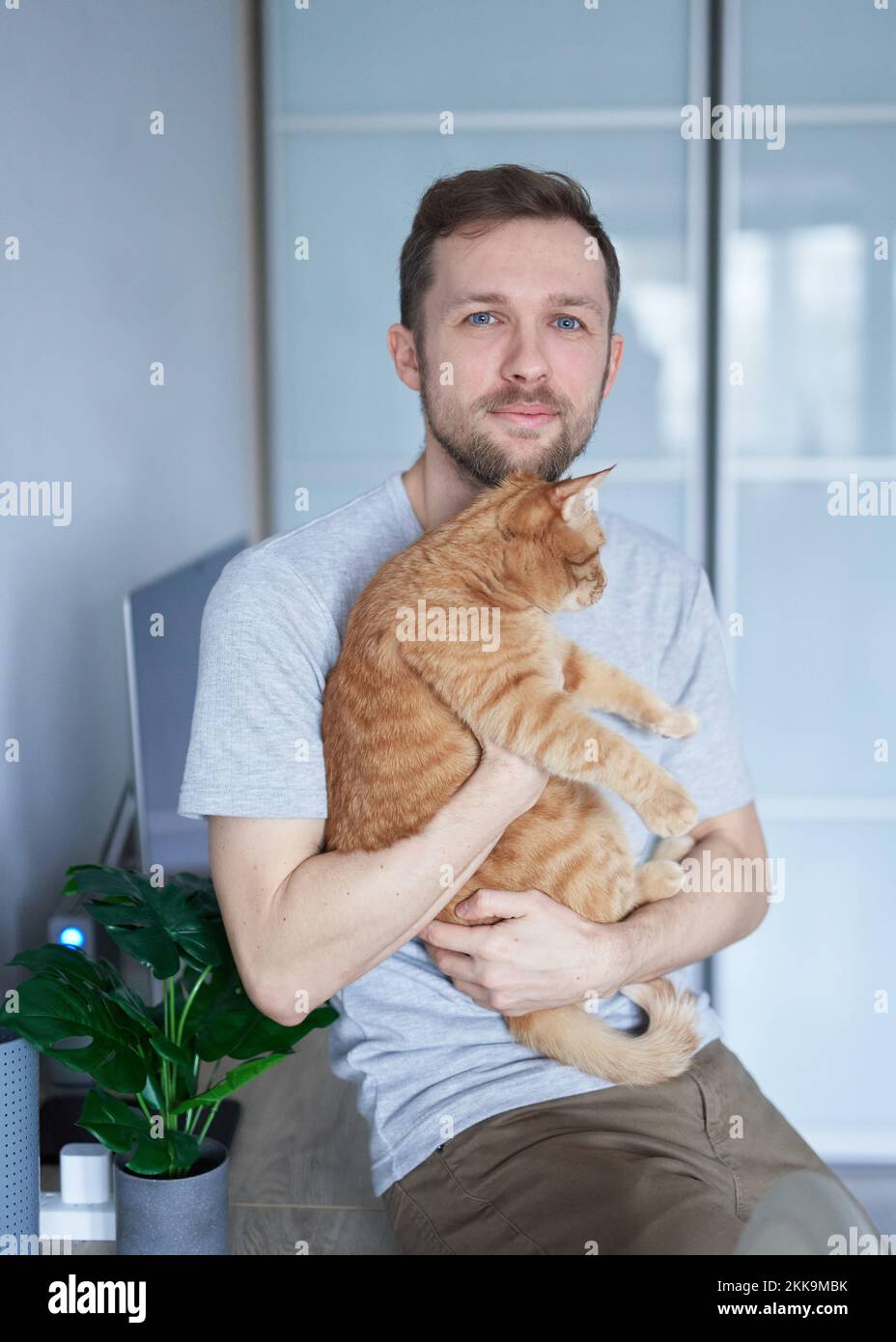 Junger Freiberufler für Software-Entwickler, männlich in grauem T-Shirt. Porträt eines fröhlichen, weißhäutigen IT-Support-Technikers am Arbeitsplatz im Heimbüro in der Nähe des Computers, der Tabby Cat in die Kamera schaut Stockfoto