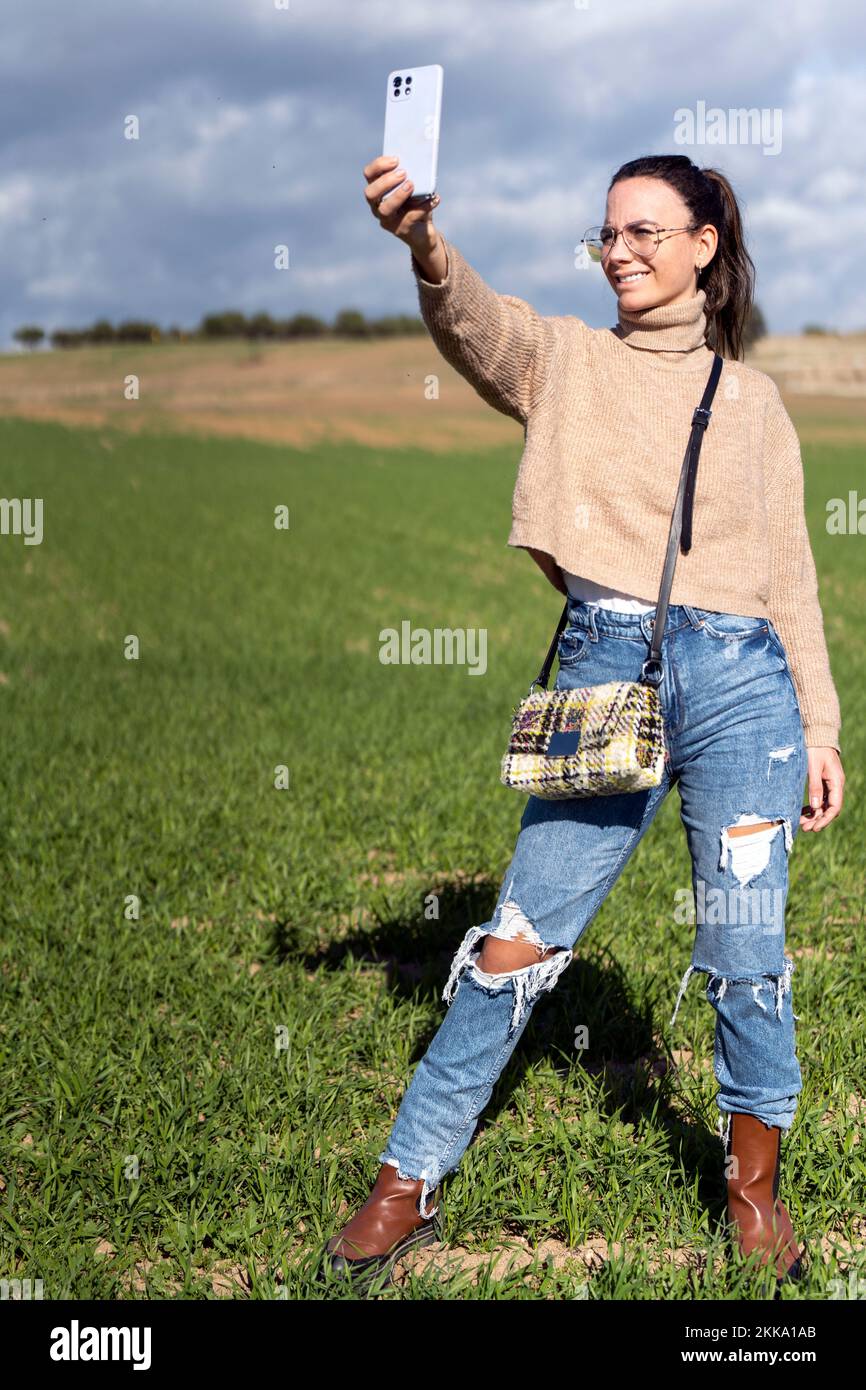 Fröhliche junge Frau in beigefarbenen Pullover-Jeans und Brille, die lächelt und Selfie auf dem Smartphone macht, während sie auf einer grünen Wiese steht Stockfoto