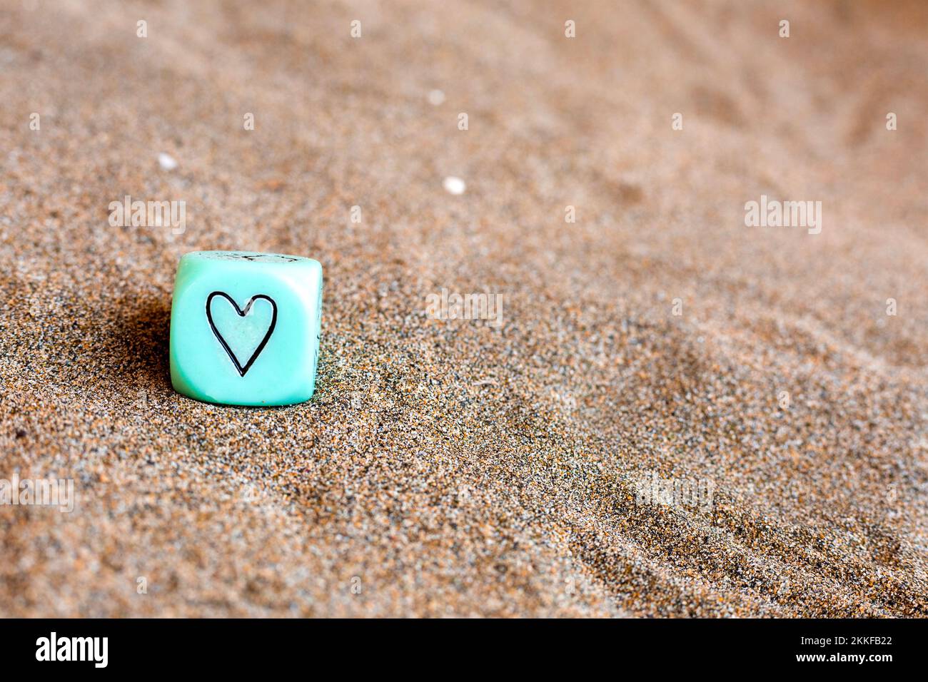 hellblauer Würfel mit Herz- und Wettersymbolen an den Seiten. Lustige Spielwürfel auf Sand. Valentinstag-Konzept. Liebessymbol. Nahaufnahme mit Kopie Stockfoto