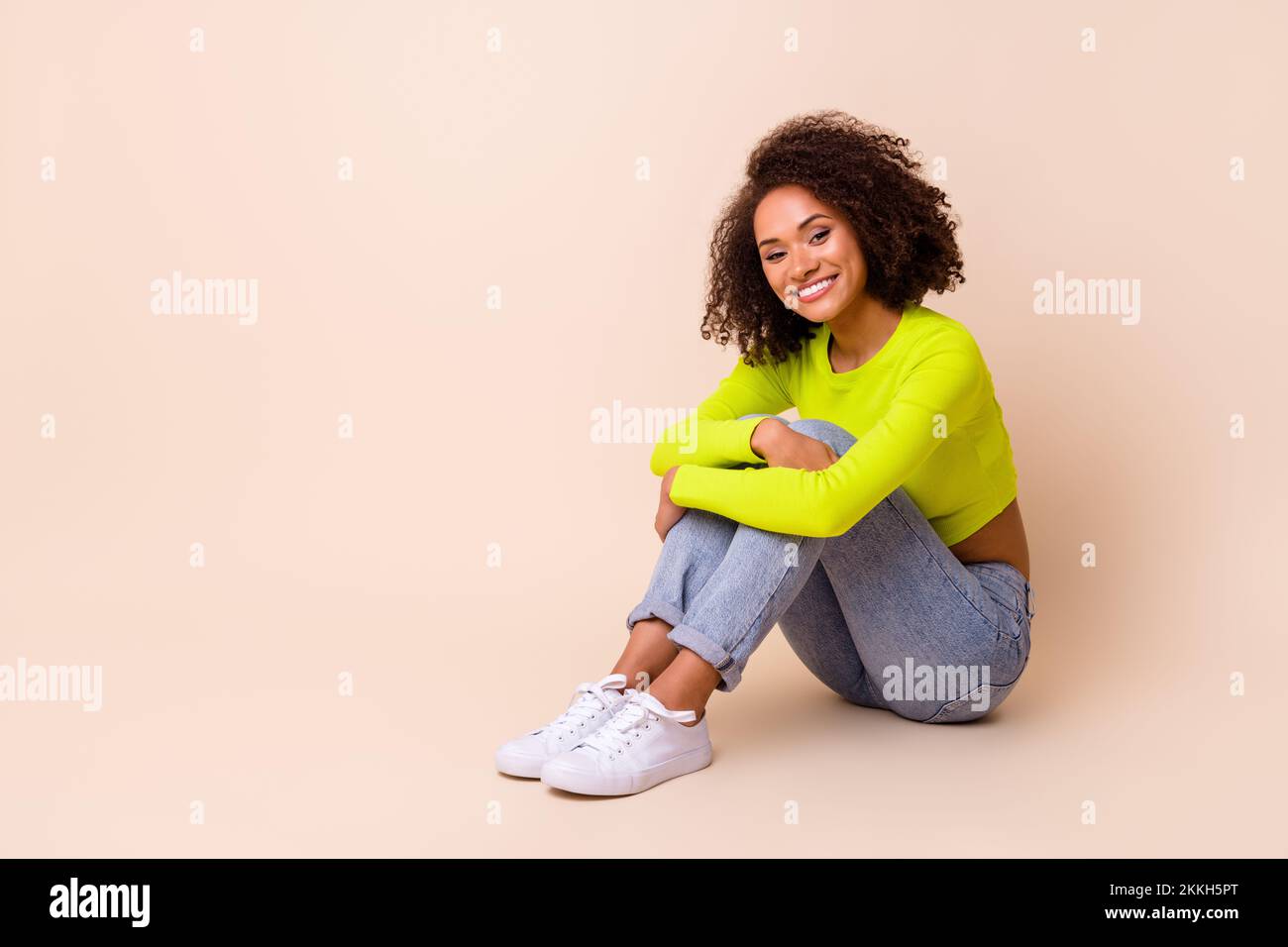 Ganzkörperfoto eines netten jungen Mädchens wellenförmiges Haar Sitz Boden Knie umarmen Zahnfleisch Lächeln Tragen Sie trendiges gelbes Outfit isoliert auf beigefarbenem Hintergrund Stockfoto