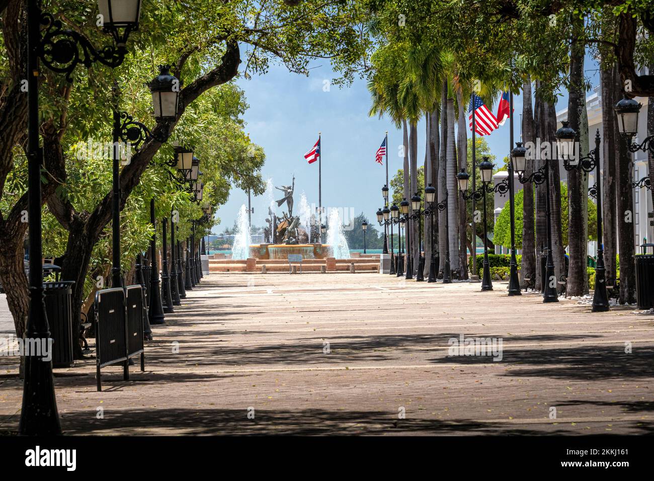 Flaggen und Bäume säumen den Paseo de la Princesa Walkway und den Raizes Fountain in Old San Juan auf der tropischen karibischen Insel Puerto Rico, USA. Stockfoto