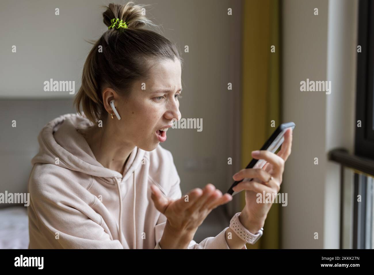 Junge kaukasische Frau mit Mobiltelefon und kabellosen Kopfhörern, Ruhe, sitzen auf dem Bett zu Hause und hören Podcast oder Clubhaus - nur Stimme Stockfoto