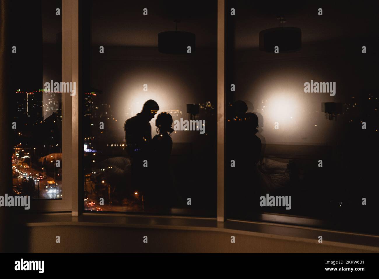 Reflektiert von liebevollem Paar, das sich auf einem Panoramafenster küsst und bei Nacht einen Blick auf die Stadt hat. Romantik-in-the-Sky-Konzept Stockfoto