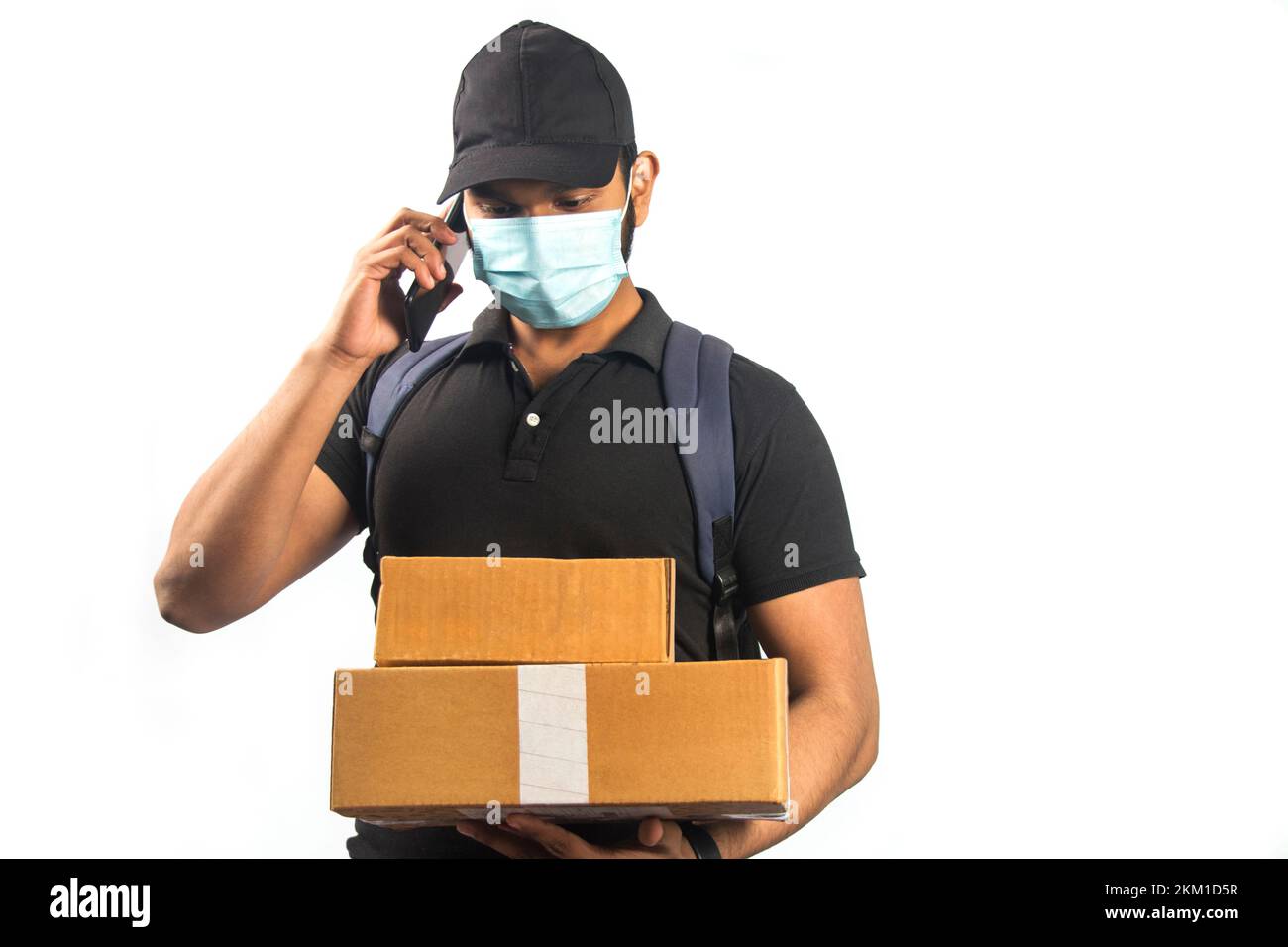 Ein glücklicher indischer Zusteller, der mit einem Smartphone spricht, und ein Paket in schwarzer Uniform Stockfoto
