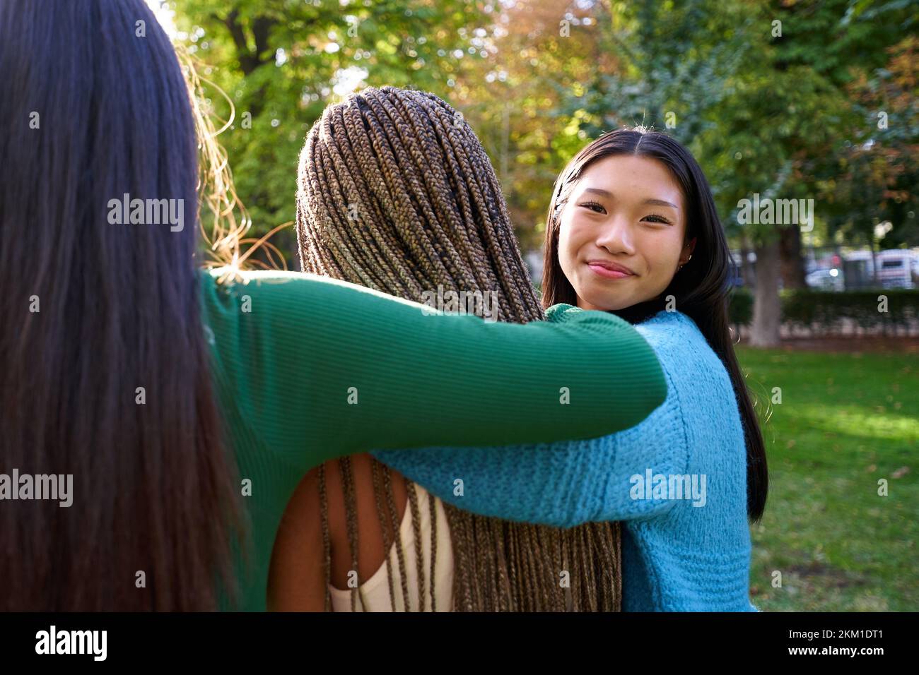 Drei Freundinnen umarmen sich in einem öffentlichen Park, während eine von ihnen lächelnd in die Kamera schaut. Stockfoto
