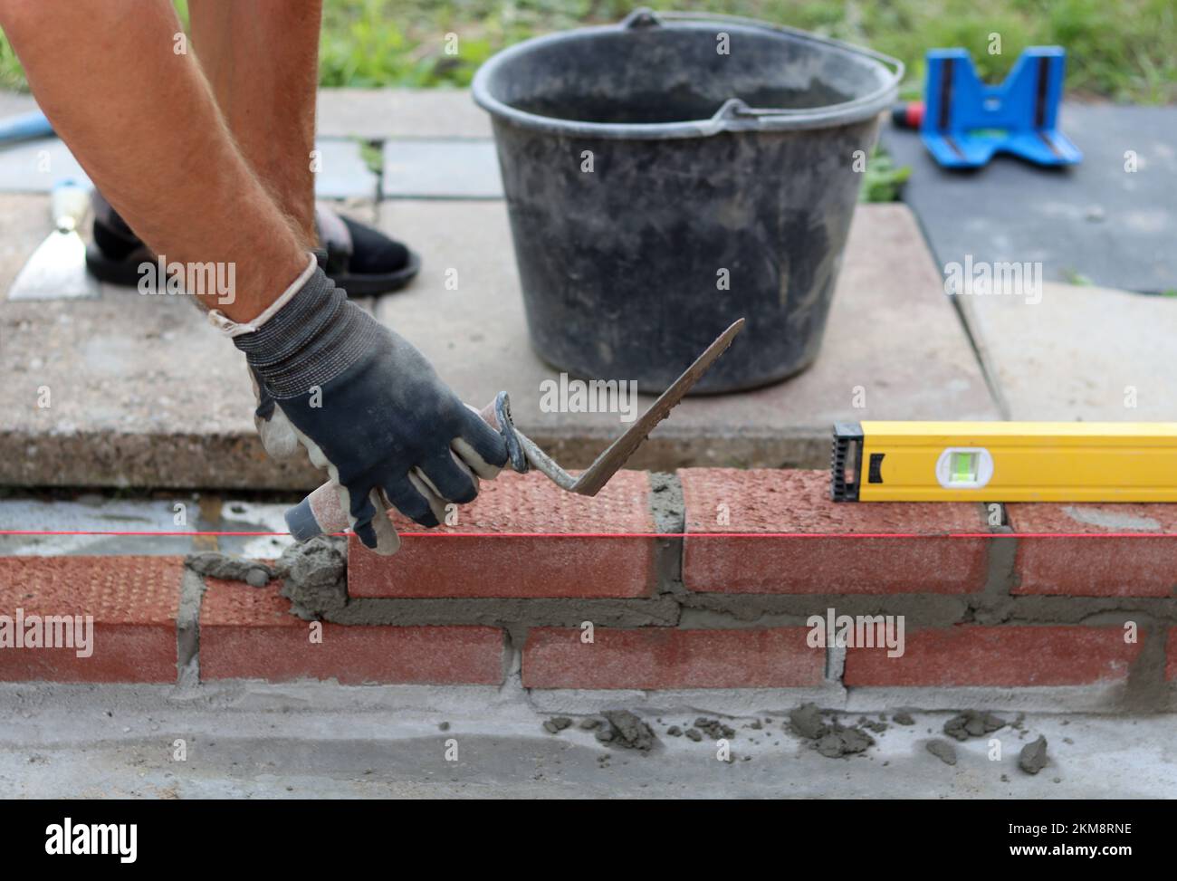 Eine Mauer aus roten Ziegeln zu bauen. Männliche Hände halten Maurerkelle und Nivelliermessgerät. Mauerwerk im Detail. Nahaufnahme der Baustelle. Stockfoto