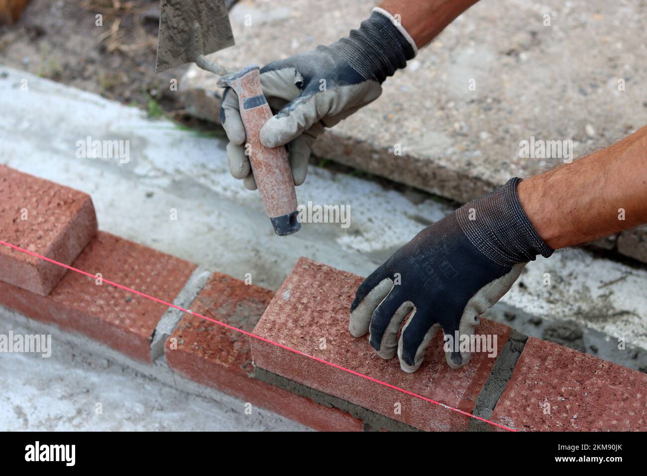 Eine Mauer aus roten Ziegeln zu bauen. Männliche Hände halten Maurerkelle und Nivelliermessgerät. Mauerwerk im Detail. Nahaufnahme der Baustelle. Stockfoto