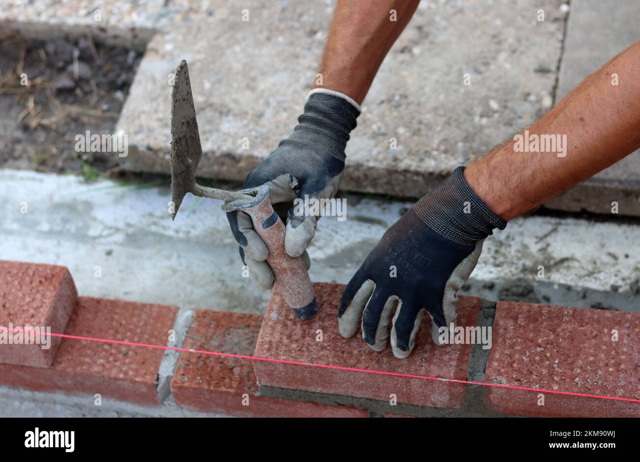 Eine Mauer aus roten Ziegeln zu bauen. Männliche Hände halten Maurerkelle und Nivelliermessgerät. Mauerwerk im Detail. Nahaufnahme der Baustelle. Stockfoto