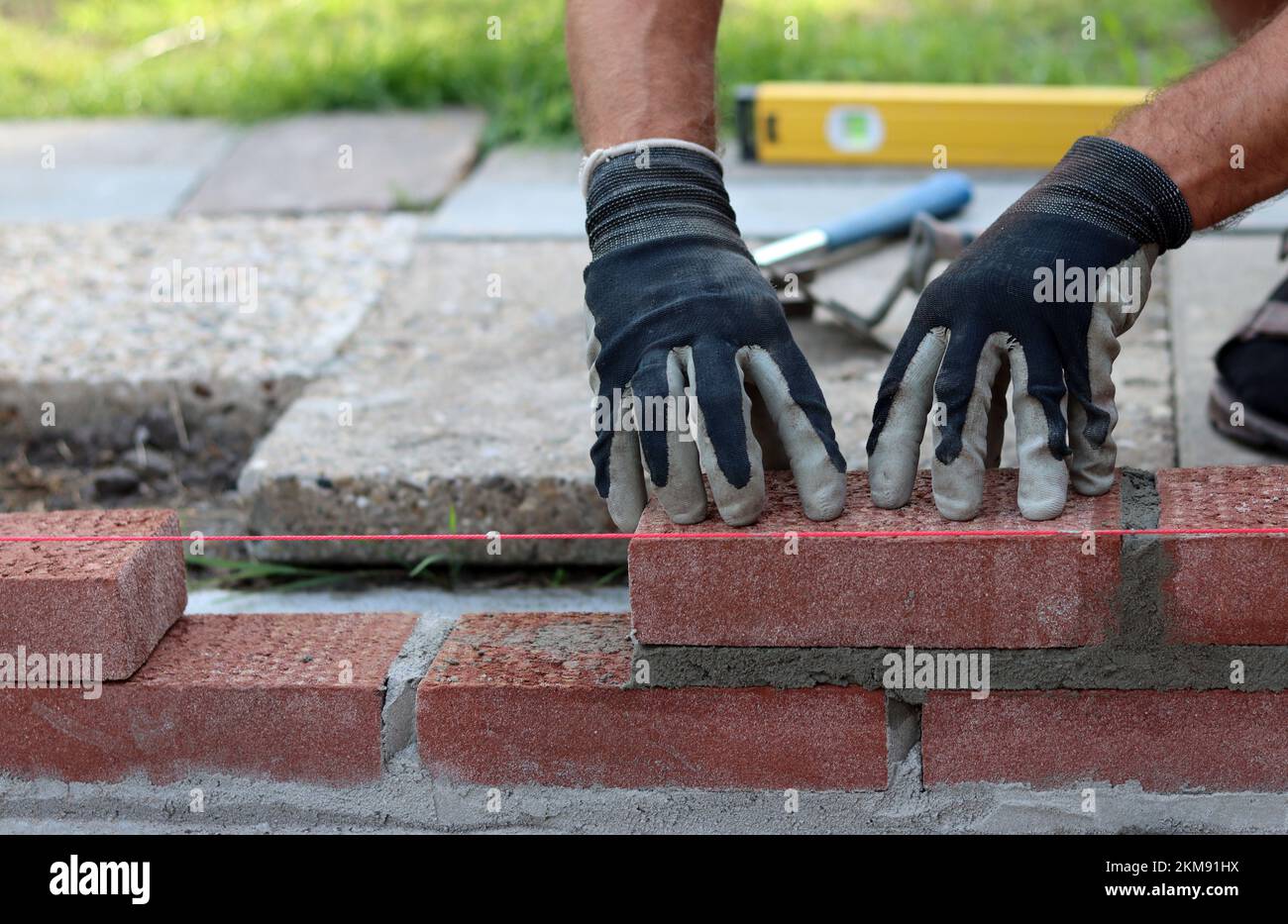 Eine Mauer aus roten Ziegeln zu bauen. Männliche Hände halten Maurerkelle und Nivelliermessgerät. Mauerwerk im Detail. Nahaufnahme der Baustelle. Stockfoto
