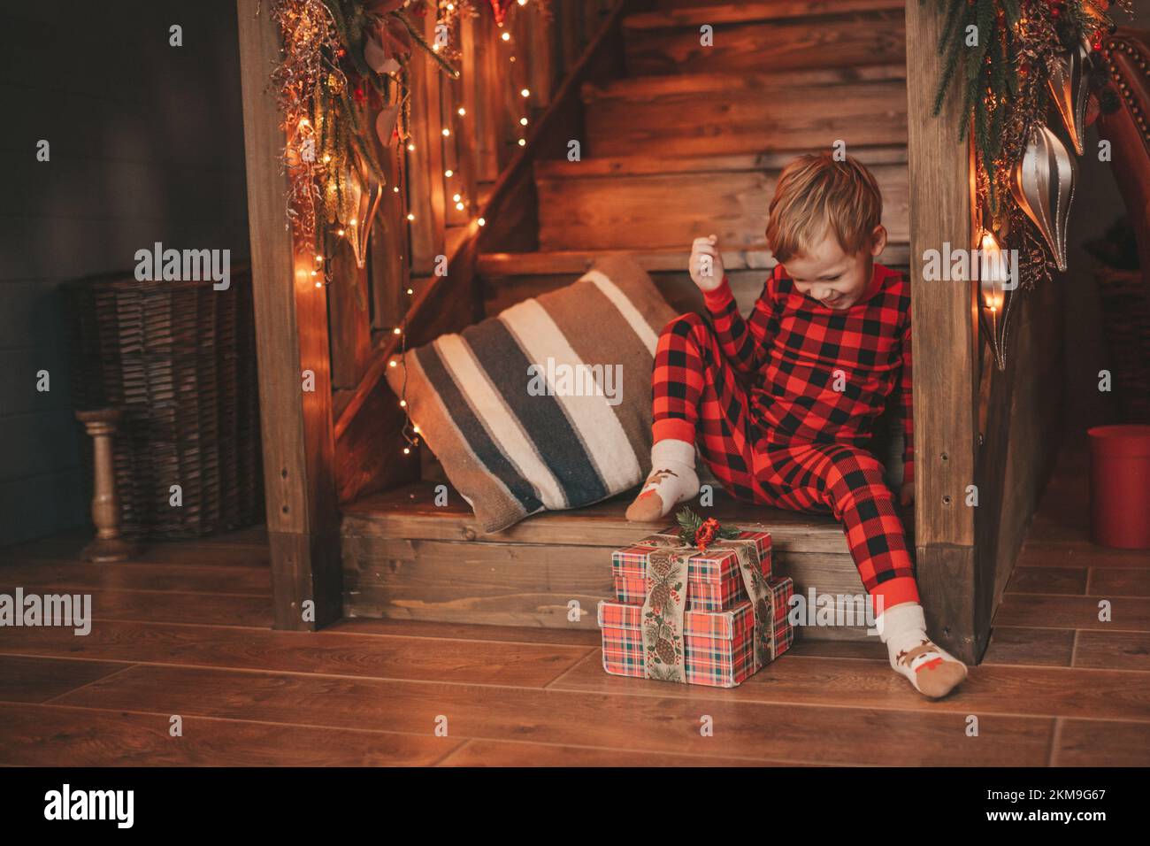 Lächeln kleines Kind aktiv mit Spaß warten auf Wunder Santa Open Geschenke. Rot karierte Nachwäsche fröhliches Kind feiert Neujahr mit Geschenk Stockfoto