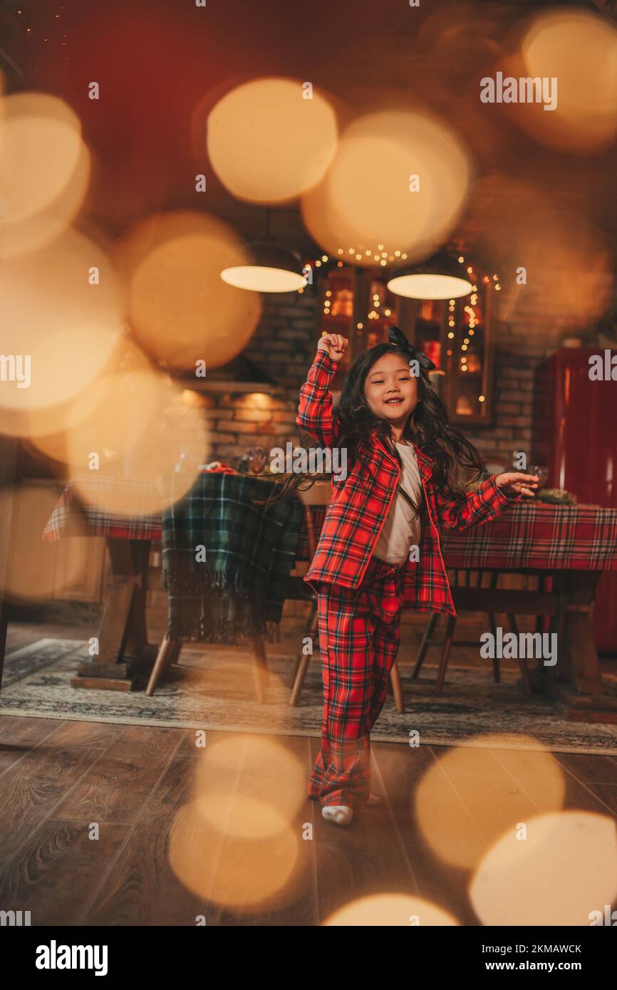 Active japan Kid Girl mit stilvollen langen Brünette Haarmodell zu Hause in Bokeh Girlanden noel posieren. Asiatische kleine Fashionista in rot karierten Pyjama tanzen Stockfoto